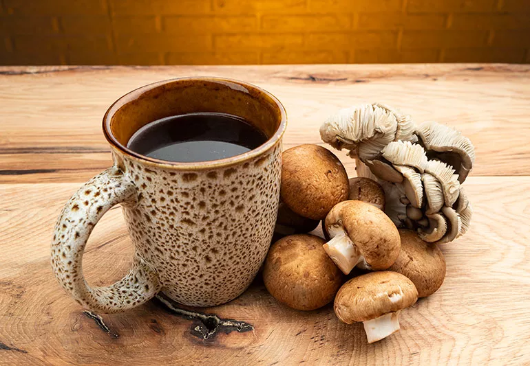 A cup of mushroom coffee surrounded by coffee beans and various mushrooms on a rustic wooden table with fresh green leaves.