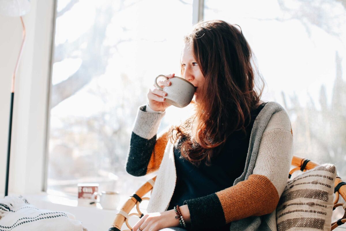 Smiling woman sipping coffee from a ceramic mug, seated in a cozy chair by a bright window.