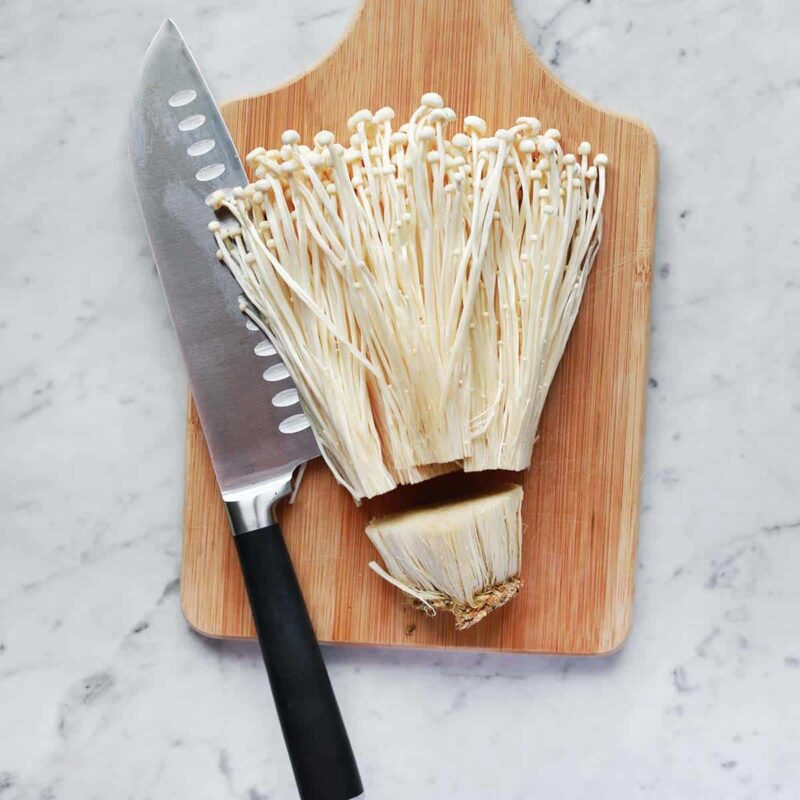 Fresh enoki mushrooms on a wooden cutting board next to a chef's knife, ready for cooking.